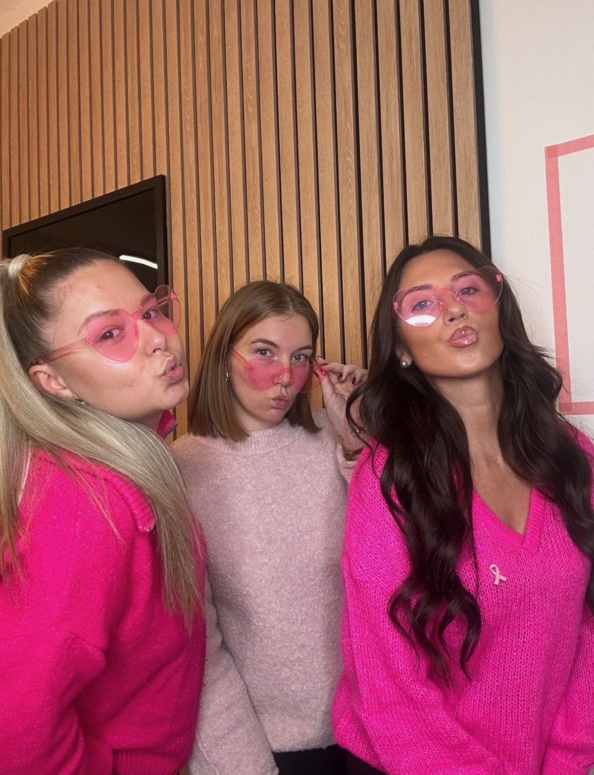 Trois filles de l’équipe en pulls roses et lunettes cœur, ruban rose épinglé