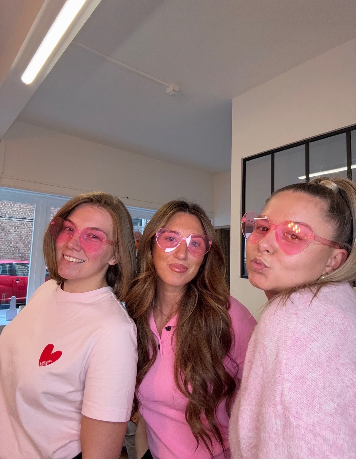Trois filles de l’équipe en rose avec des lunettes cœur, dans le salon